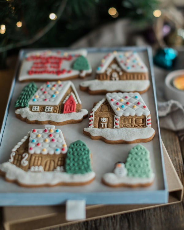 Honeywell Bakes Personalised Christmas Village Biscuit Gift - Six decorated gingerbread house cookies arranged on a light blue tray, with festive blurred holiday lights and ornaments in the background.