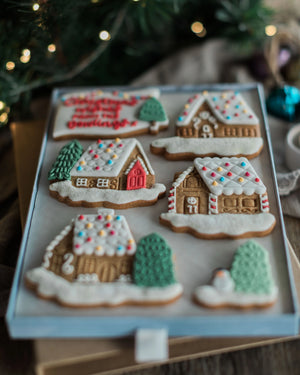 Honeywell Bakes Personalised Christmas Village Biscuit Gift - A tray of six decorated gingerbread cookies shaped like houses and trees, arranged on a light blue board with a festive holiday background and blurred lights.