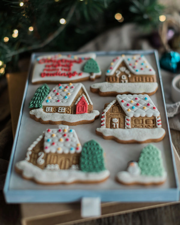 Honeywell Bakes Personalised Christmas Village Biscuit Gift - A tray of six decorated gingerbread cookies shaped like houses and trees, arranged on a light blue board with a festive holiday background and blurred lights.