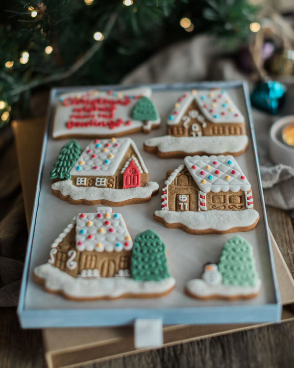 Honeywell Bakes Personalised Christmas Village Biscuit Gift - A set of decorated gingerbread cookies shaped like holiday houses and trees, arranged in a light blue tray on a wooden surface with festive blurred lights and greenery in the background.