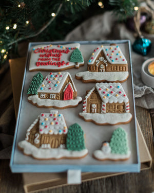 Honeywell Bakes Personalised Christmas Village Biscuit Gift - A tray of decorated gingerbread cookies shaped like winter houses and trees, photographed on a rustic wooden surface with holiday lights and greenery in the background.
