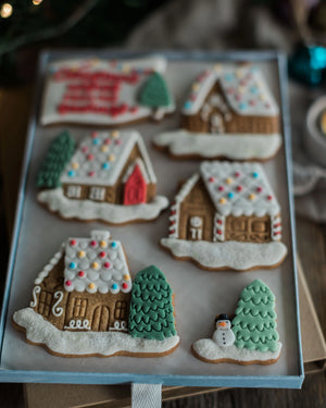 Honeywell Bakes Personalised Christmas Village Biscuit Gift - Six decorated gingerbread cookies shaped like winter houses, trees, and a snowman, arranged on a light blue tray against a softly blurred holiday background.