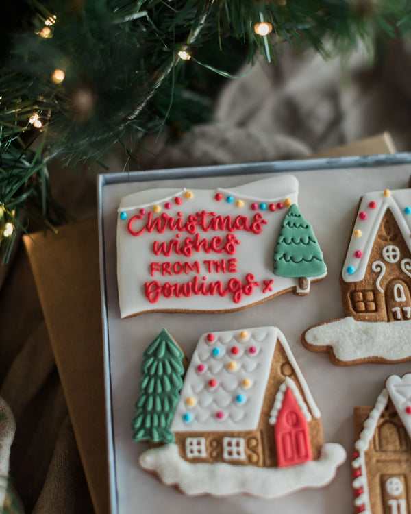 Honeywell Bakes Personalised Christmas Village Biscuit Gift - A close-up of decorated Christmas gingerbread cookies in a tray, including a banner cookie with the text 'Christmas wishes FROM THE Dowlings' and snow-covered house cookies, with blurred evergreen branches and warm string lights in the background.