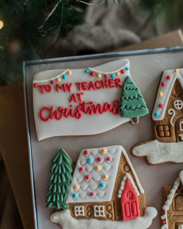 Honeywell Bakes Personalised Christmas Village Biscuit Gift - Decorated gingerbread cookies shaped like a house and a waving banner, with one cookie reading 'TO MY TEACHER AT Christmas' in red icing, displayed on a baking sheet with a blurred holiday background.