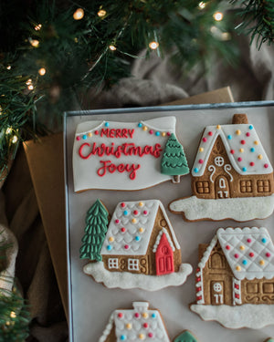 Honeywell Bakes Personalised Christmas Village Biscuit Gift - A tray of decorated Christmas gingerbread cookies, including a sign that reads 'Merry Christmas Joy' and several gingerbread houses with colourful icing snow and accents, styled under warm holiday lights with pine branches in the background.