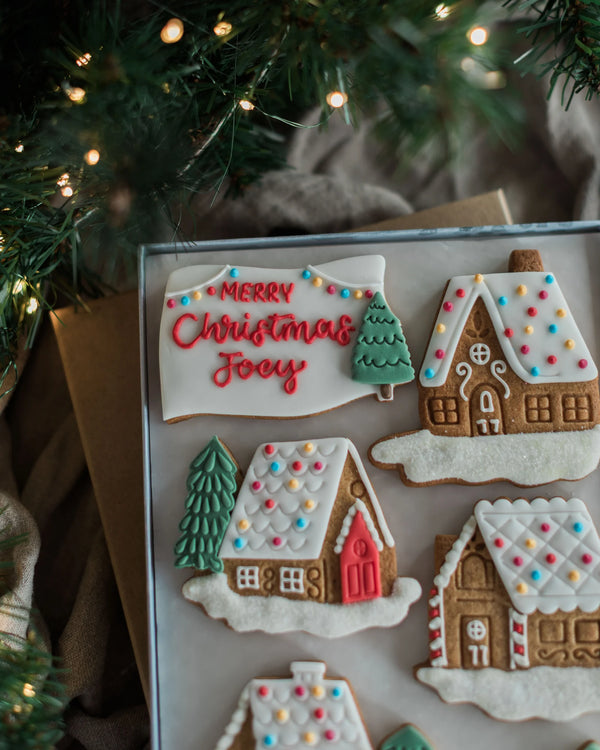Honeywell Bakes Personalised Christmas Village Biscuit Gift - A tray of decorated Christmas gingerbread cookies, including a sign that reads 'Merry Christmas Joy' and several gingerbread houses with colourful icing snow and accents, styled under warm holiday lights with pine branches in the background.