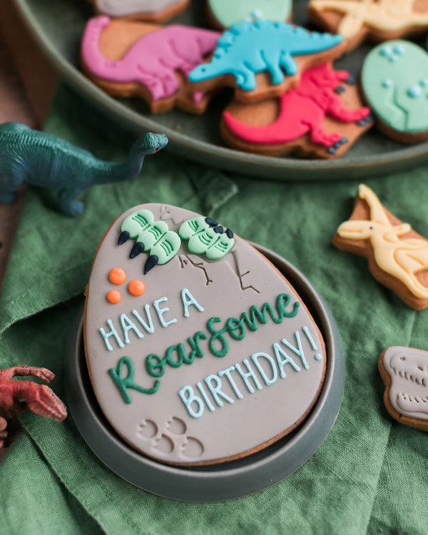 Honeywell Bakes Personalised Dinosaur Egg Biscuit - Dinosaur-themed cookies with a 'Have a Roarsome Birthday!' cookie on a green surface.