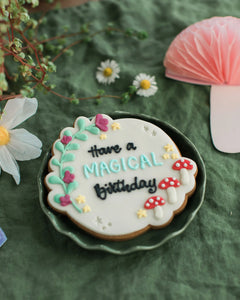 Honeywell Bakes Personalised Fairy Circle Biscuit - Decorative cookie with 'Have a Magical Birthday' text on a green surface with flowers and a mushroom.