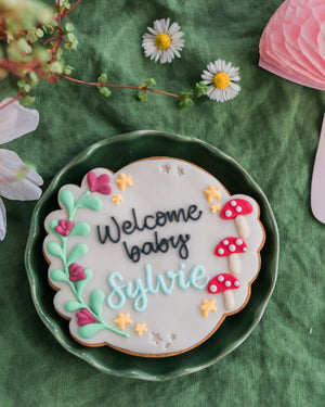 Honeywell Bakes Personalised Fairy Circle Biscuit - Decorative cookie with 'Welcome baby Sylvie' text on a green plate with flowers in the background
