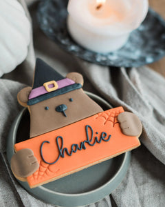 Honeywell Bakes Personalised Halloween Biscuit Bear - Cookie shaped like a bear wearing a witch hat, holding an orange sign with 'Charlie' on it, placed on a gray surface with a candle in the background.