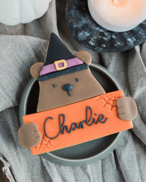 Honeywell Bakes Personalised Halloween Biscuit Bear - Cookie shaped like a bear wearing a witch hat, holding an orange sign with 'Charlie' on it, placed on a gray surface with a candle in the background.