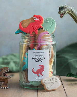Honeywell Bakes Personalised Jar of Dinosaur Biscuits - Jar of dinosaur-shaped cookies with a label for a dinosaur party on a wooden surface.