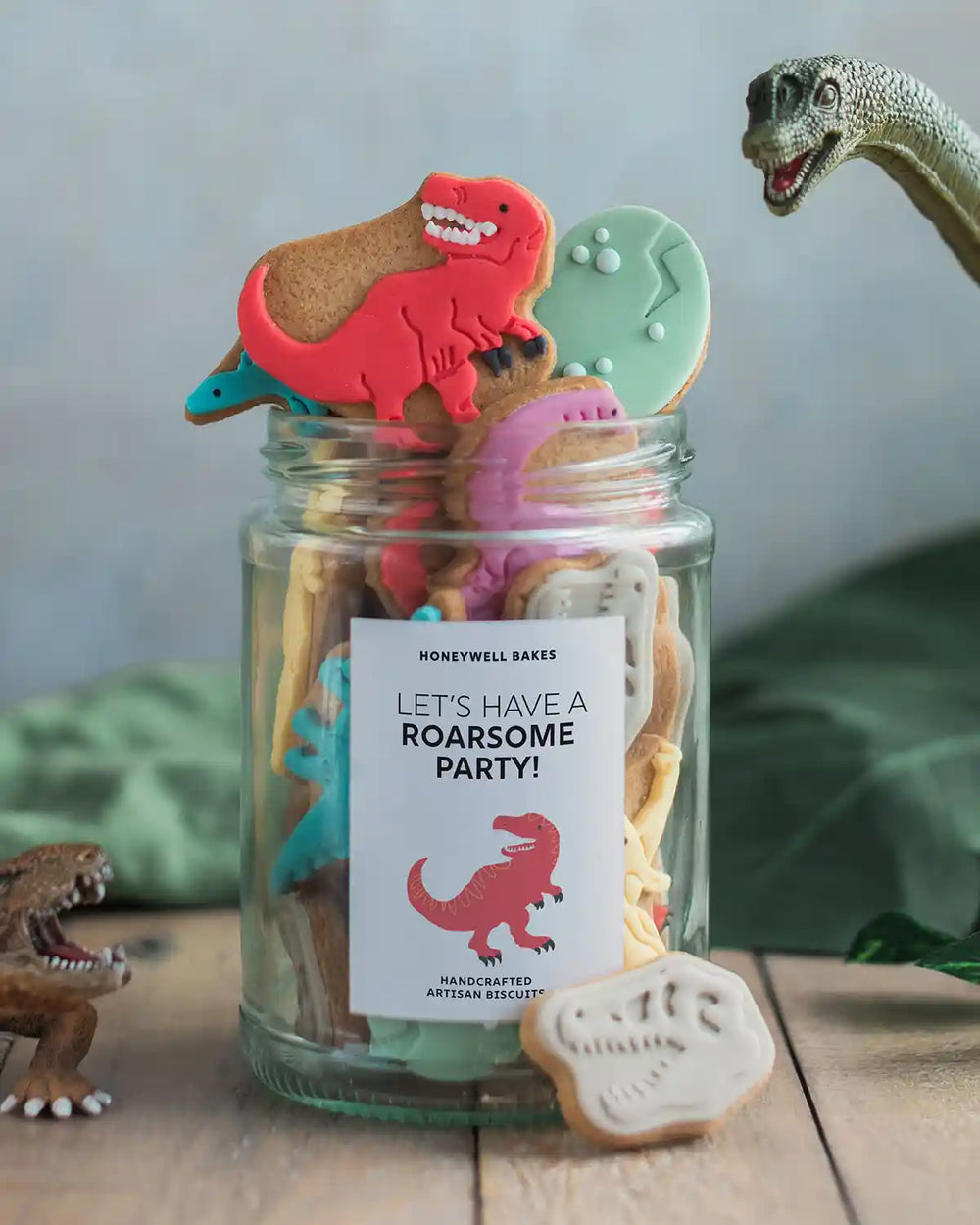 Honeywell Bakes Personalised Jar of Dinosaur Biscuits - Jar of dinosaur-shaped cookies with a 'Let's have a roarsome party!' label, surrounded by small dinosaur figurines.