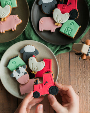 Honeywell Bakes Personalised Jar of Farm Animal Biscuits - Farm-themed cookies shaped like animals and vehicles on plates with a hand holding a red truck cookie.