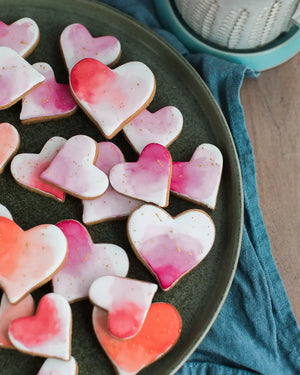 Honeywell Bakes Personalised Jar of Heart Biscuits - Heart-shaped cookies with pink and red watercolour design on a dark plate.