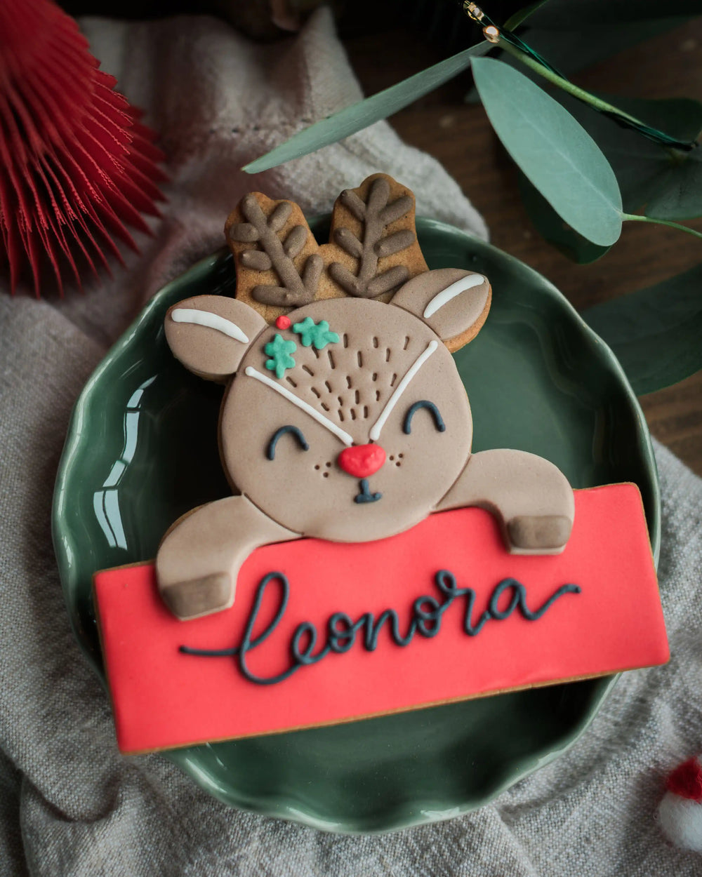 A Personalised Reindeer Biscuit, personalised with the name 'Leonora' in black royal icing, styled on a green plate with holiday decor in the background.