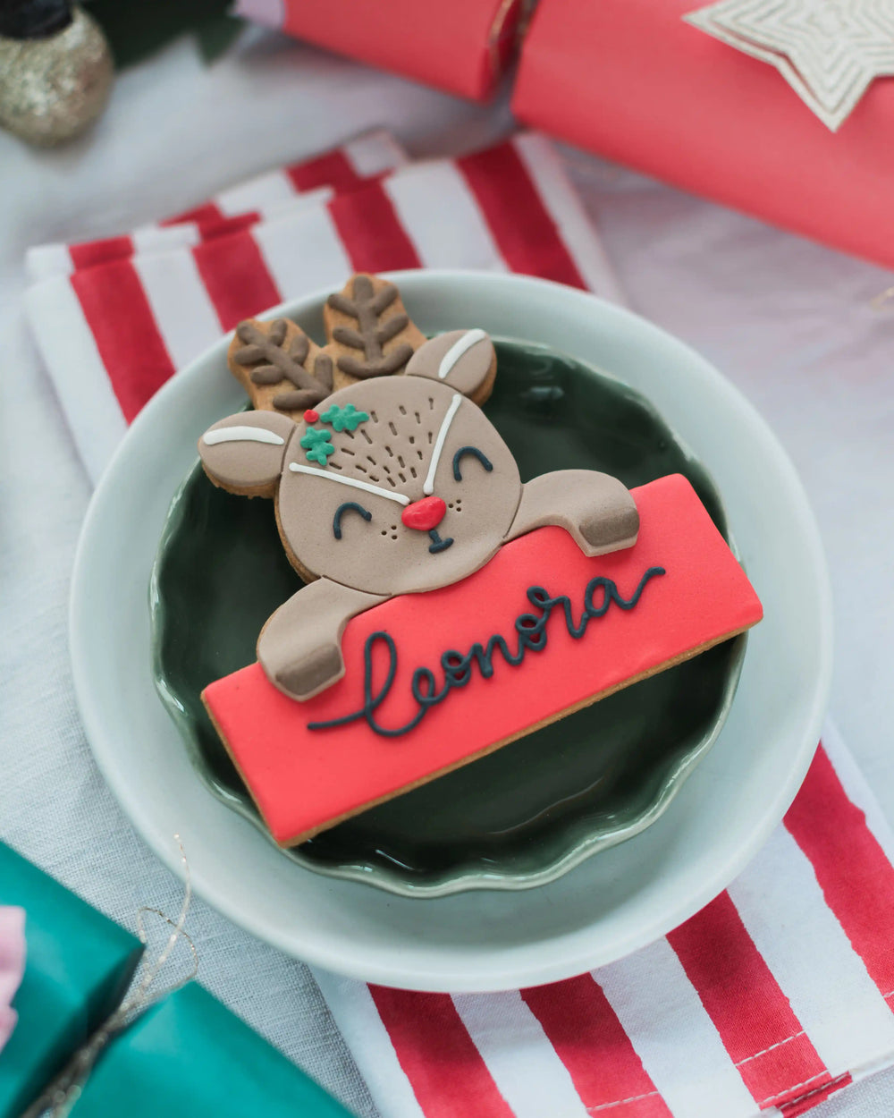 A Personalised Reindeer Biscuit, personalised with the name 'Leonora' in black royal icing, styled on a green plate with holiday decor in the background.