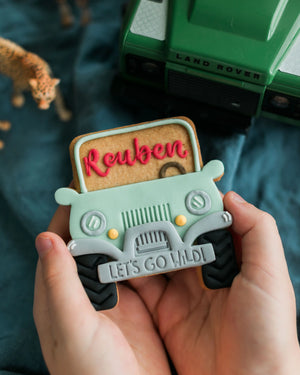 Honeywell Bakes Personalised Safari Car Biscuit - Hand holding a truck biscuit with 'Reuben' and 'Let's Go Wild!' text, Land Rover in the background