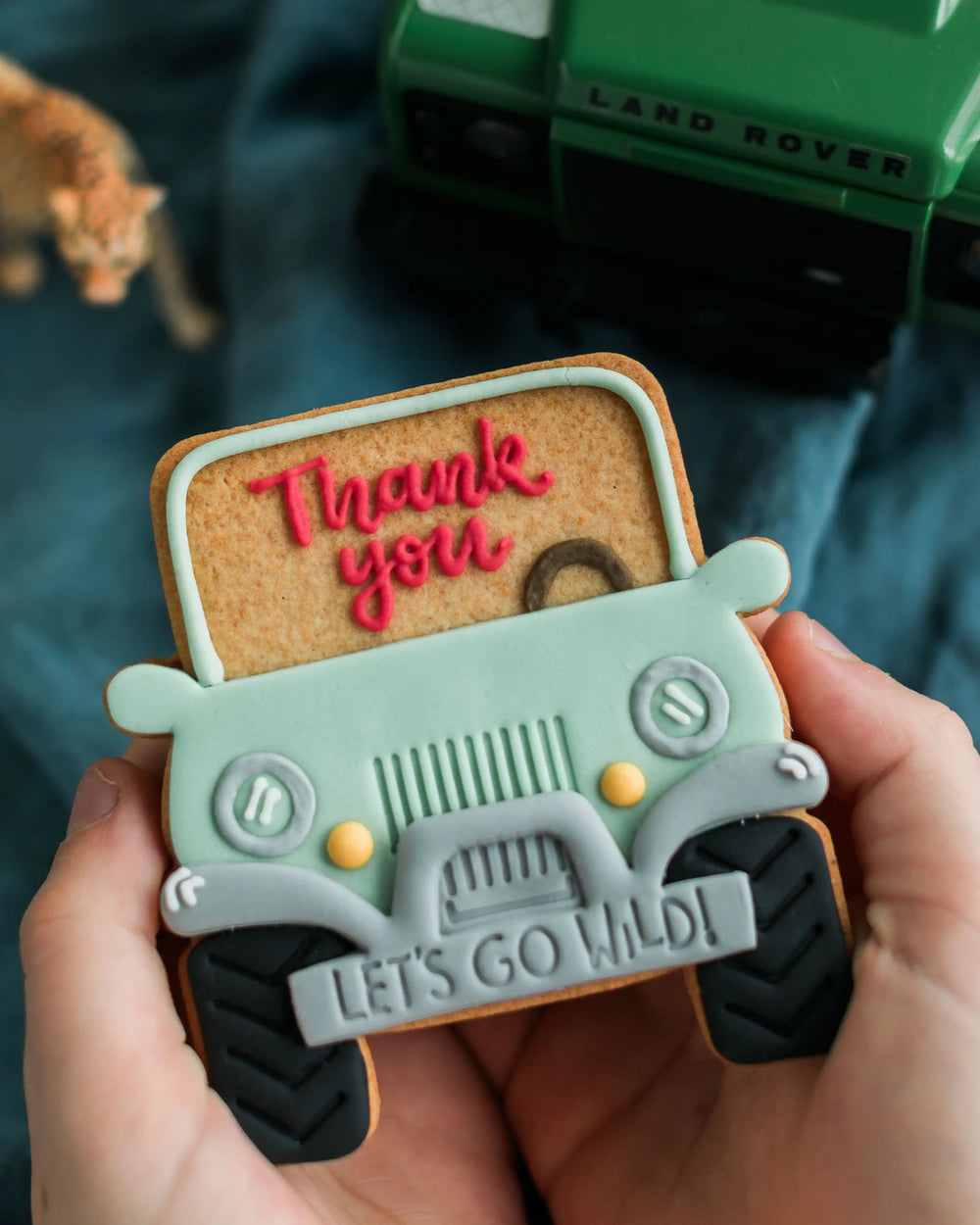 Honeywell Bakes Personalised Safari Car Biscuit - Cookie-shaped Jeep with 'Thank you' and 'Let's go wild!' text, held by hands with a blurred Land Rover in the background.