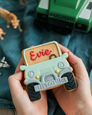 Honeywell Bakes Personalised Safari Car Biscuit - Cookie with 'Evie' and 'Let's Go Wild!' text, held by hands on a blue fabric background with toy cars.