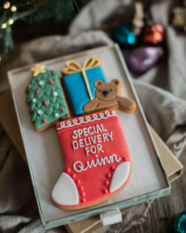 Personalised Stocking Full of Christmas Gift Set - A set of three decorated Christmas sugar cookies displayed in a clear glass tray. The cookies include a green Christmas tree with red and white ornaments, a blue gift box with a gold bow, a brown teddy bear, and a red stocking with white 'SPECIAL DELIVERY FOR Quinn' icing.