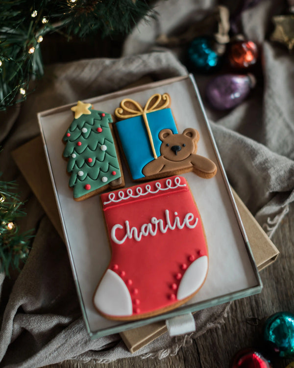 Personalised Stocking Full of Christmas Gift Set - A set of decorated Christmas-themed sugar cookies in a clear gift box, including a green Christmas tree, a blue gift box, a brown teddy bear, and a red stocking with the name 'Charlie' in white icing. The box is styled on a rustic wooden surface with a beige cloth, holiday ornaments, and evergreen branches.