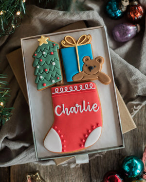 Personalised Stocking Full of Christmas Gift Set - A boxed holiday cookie shaped like a Christmas stocking with the name 'Charlie' in white icing, accompanied by a green tree, blue gift box, and brown teddy bear, displayed on a rustic fabric surface with Christmas ornaments around it.