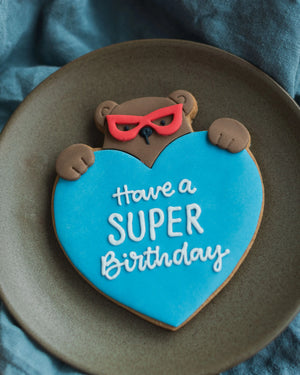 Honeywell Bakes Personalised Super Bear Hug Biscuit - A hand iced biscuit bear wearing a super hero mask and holding a love heart personalised with text 'Have a super birthday'