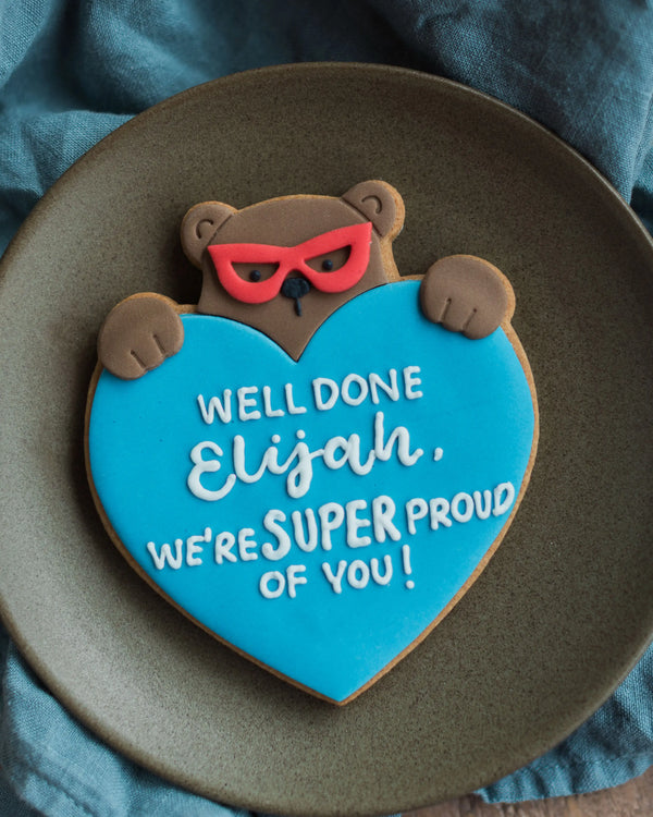 Honeywell Bakes Personalised Super Bear Hug Biscuit - A hand iced biscuit bear wearing a super hero mask and holding a love heart personalised with text Well Done We’re Super Proud of you!