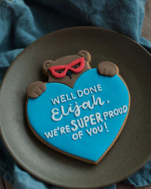 Honeywell Bakes Personalised Super Bear Hug Biscuit - A hand iced biscuit bear wearing a super hero mask and holding a love heart personalised with text Well Done We’re Super Proud of you!