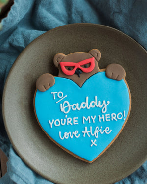 Honeywell Bakes Personalised Super Bear Hug Biscuit - A hand iced biscuit bear wearing a super hero mask and holding a love heart personalised with text To Daddy You’re My Hero Love Alfie x