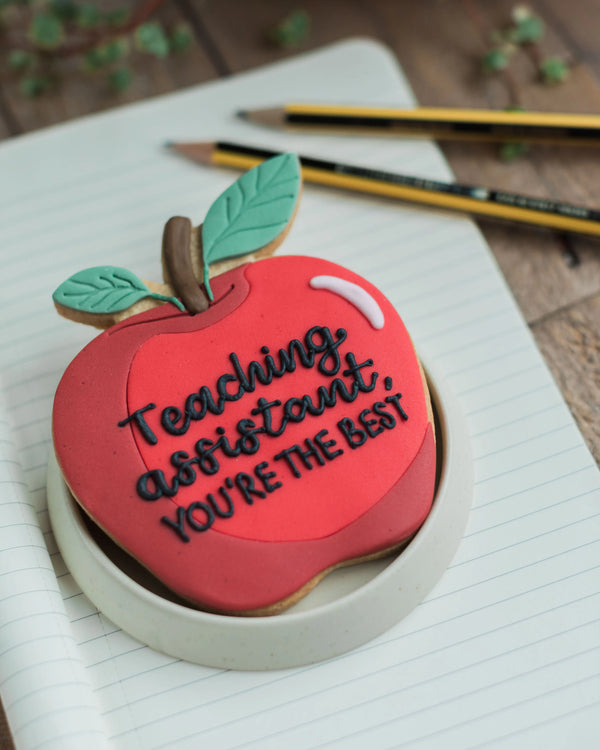 Honeywell Bakes Personalised Thank You Teacher Apple Biscuit - Red apple-shaped cookie with 'Teaching assistant, you're the best' text on a notebook with pencils.