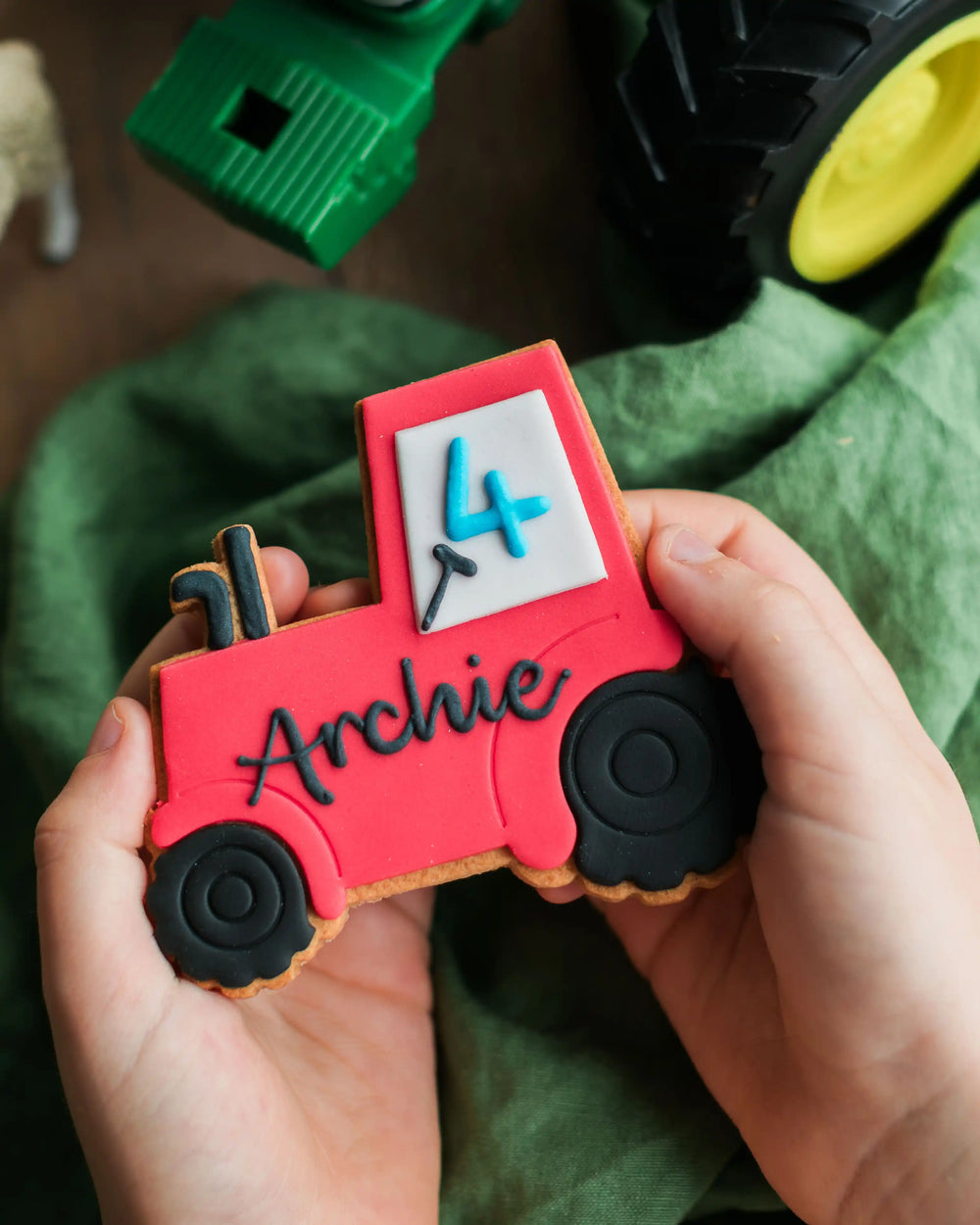 Honeywell Bakes Personalised Tractor Biscuit - Red tractor cookie with 'Archie' on it held by hands against a green fabric background.