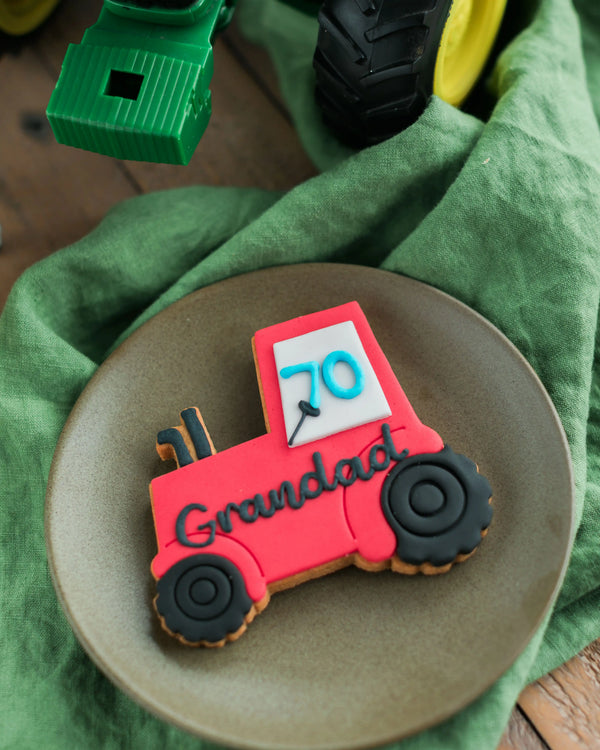 Honeywell Bakes Personalised Tractor Biscuit - Decorative cookie shaped like a red tractor with '70 Grandad' on a green cloth background