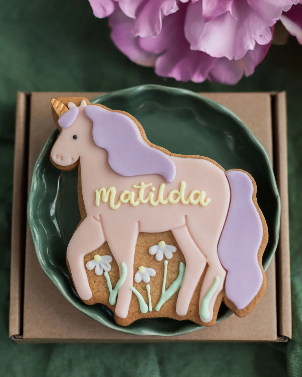 Honeywell Bakes Personalised Unicorn Biscuit - Unicorn-shaped cookie with 'Matilda' on a green plate, surrounded by pink flowers.