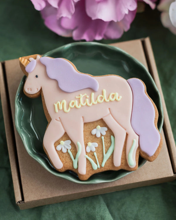 Honeywell Bakes Personalised Unicorn Biscuit - Unicorn-shaped cookie with 'Matilda' on a green plate, surrounded by pink flowers.