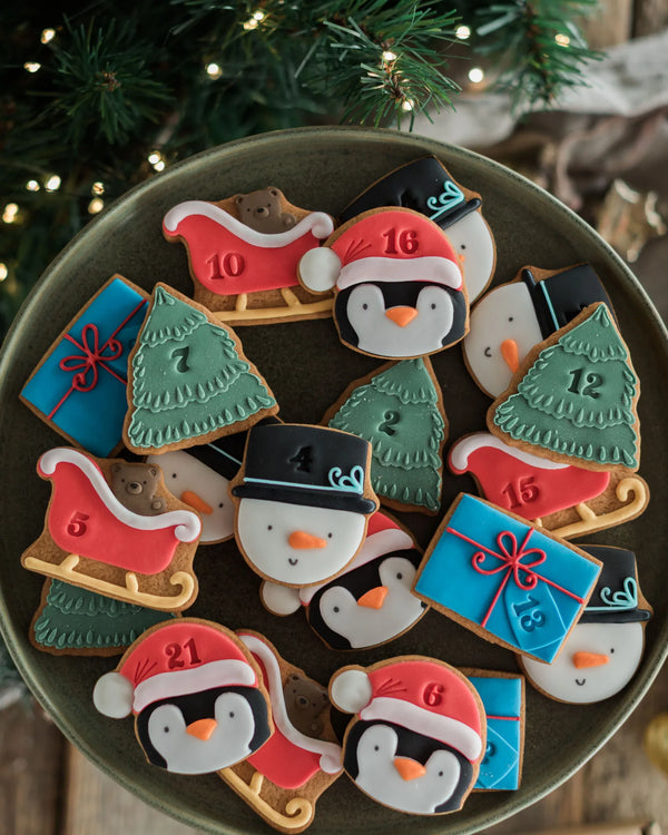 Honeywell Bakes Personalised Christmas Advent Calendar Biscuit Jar - A tray of decorated Christmas cookies shaped like penguins, sledding boats, Christmas trees, and gift boxes, arranged on a dark round plate with a festive holiday background.