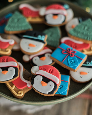 Honeywell Bakes Personalised Christmas Advent Calendar Biscuit Jar - A close-up of assorted holiday sugar cookies shaped like penguins, snowmen, Santa Claus, Christmas trees, and wrapped gifts, arranged on a dark plate.