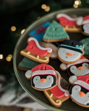Honeywell Bakes Personalised Christmas Advent Calendar Biscuit Jar - A close-up of assorted Christmas-themed iced cookies arranged on a dark green plate, including penguins in Santa hats, snowman faces, Christmas trees, and sleighs, with a blurred holiday background and warm bokeh lights.