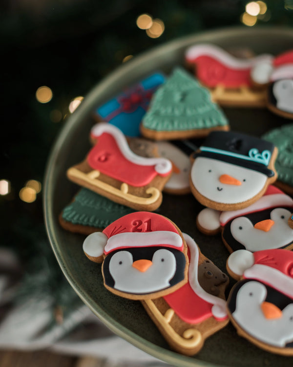 Honeywell Bakes Personalised Christmas Advent Calendar Biscuit Jar - A close-up of assorted Christmas-themed iced cookies arranged on a dark green plate, including penguins in Santa hats, snowman faces, Christmas trees, and sleighs, with a blurred holiday background and warm bokeh lights.