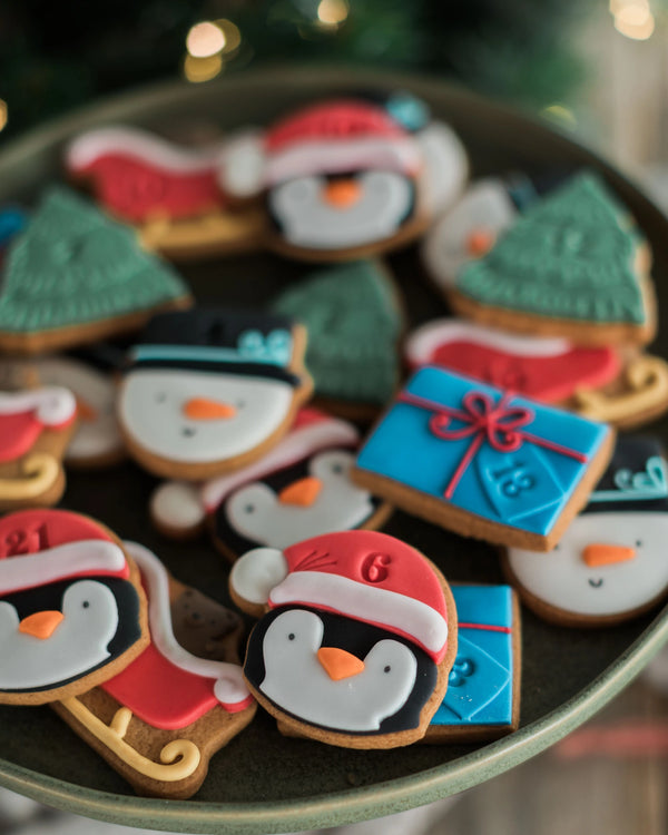 Honeywell Bakes Personalised Christmas Advent Calendar Biscuit Jar - A close-up of decorated holiday cookies shaped like penguins in Santa hats, snowmen, Christmas trees, and a blue gift box, arranged on a dark plate with a softly blurred festive background.