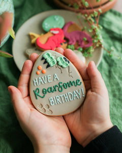 Honeywell Bakes Personalised Dinosaur Egg Biscuit - Cookie with 'Have a Roarsome Birthday!' text held by hands, with dinosaur-themed cookies in the background.