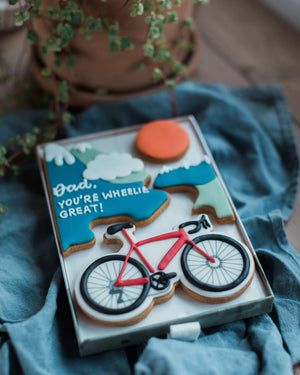 Honeywell Bakes Personalised Medium Bicycle Biscuit Gift - Bike-themed biscuits in a box with decorative elements on a blue cloth.