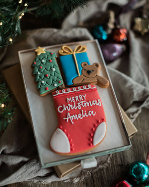 Personalised Stocking Full of Christmas Gift Set - A set of three intricately decorated Christmas sugar cookies displayed in a clear gift box on a rustic wooden surface. The cookies include a green Christmas tree with a gold star, a blue gift box with a gold bow, a brown teddy bear, and a red Christmas stocking with white 'Merry Christmas Amelia' icing.