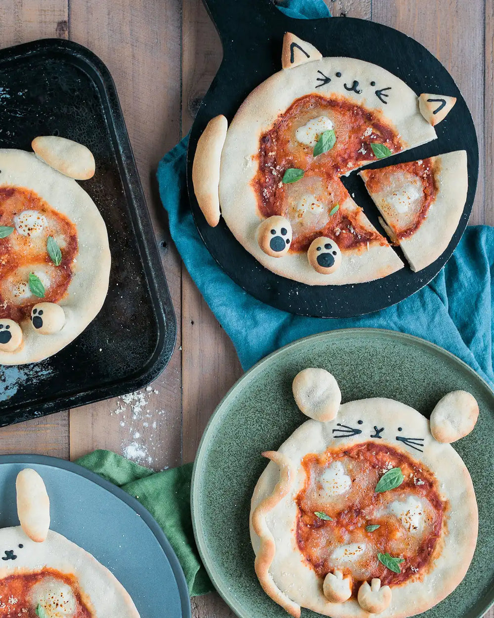 Honeywell Bakes Pizza Pet Baking Kit - Small pizzas shaped like cats on a wooden table with a blue towel underneath.