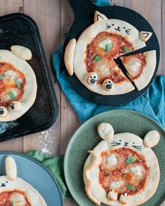 Honeywell Bakes Pizza Pet Baking Kit - Small pizzas shaped like cats on a wooden table with a blue towel underneath.