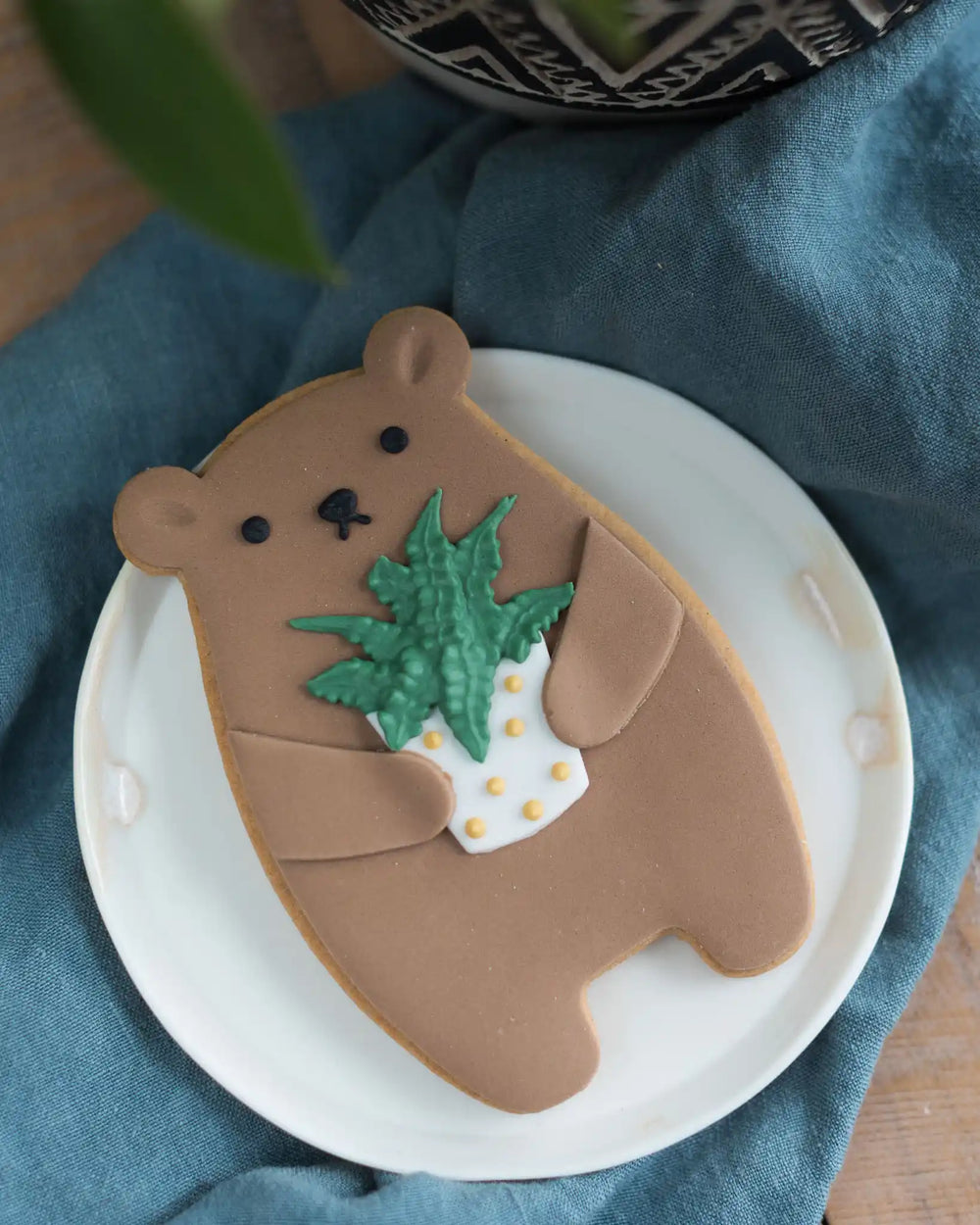 Honeywell Bakes Plant Biscuit Bear - Bear-shaped cookie holding a small plant on a white plate with a blue cloth background