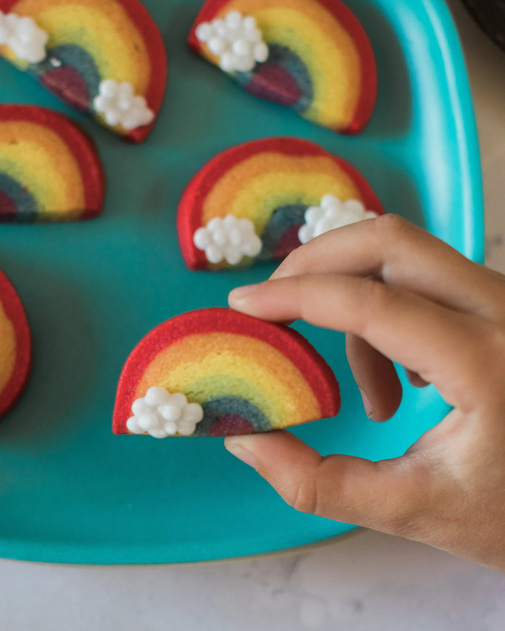 Honeywell Bakes Rainbow Biscuit Baking Kit - Hand holding a rainbow-shaped cookie with white clouds on a blue tray.