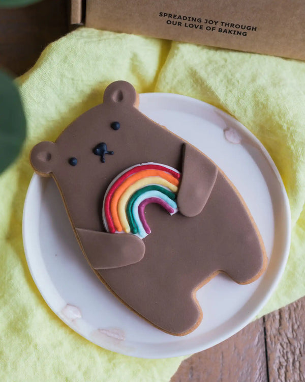 Honeywell Bakes Rainbow Biscuit Bear - Cookie shaped like a bear with a rainbow on a white plate, placed on a yellow cloth with a wooden surface underneath.