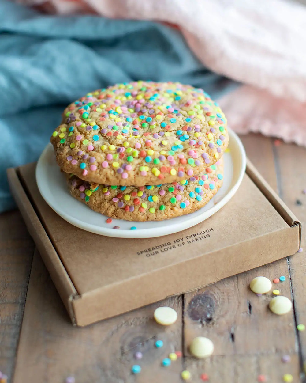 Honeywell Bakes Rainbow Sprinkle Cookie Kit - Two cookies with colourful sprinkles on a white plate on top of a brown box on a wooden surface.